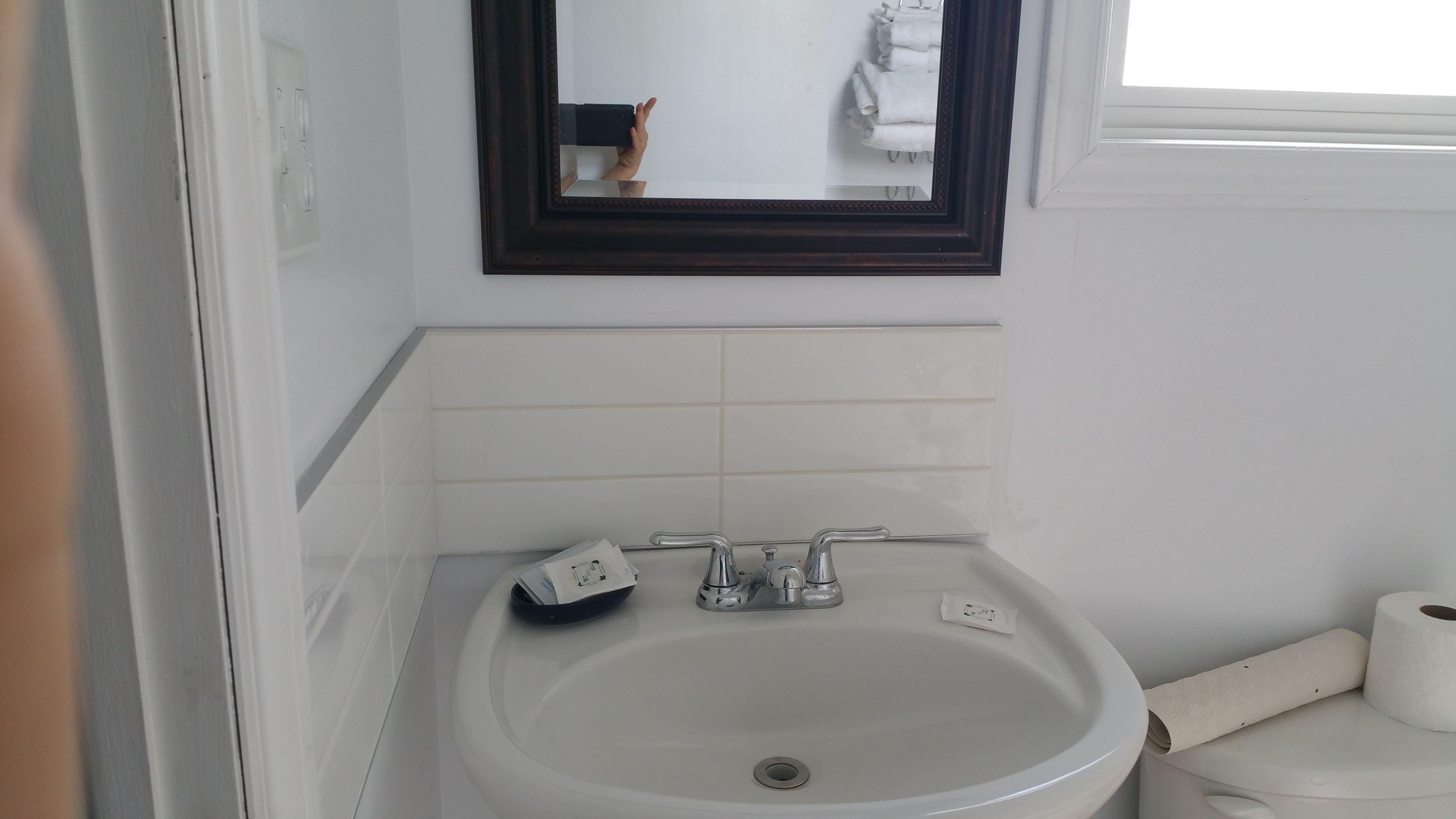 Un baño con un lavamanos blanco, dos grifos, un espejo sobre el lavamanos y una ventana a la derecha.