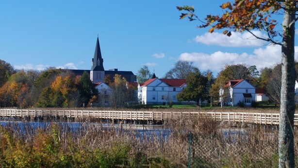 Vista a la orilla del río con una iglesia, edificios residenciales y un puente de madera.