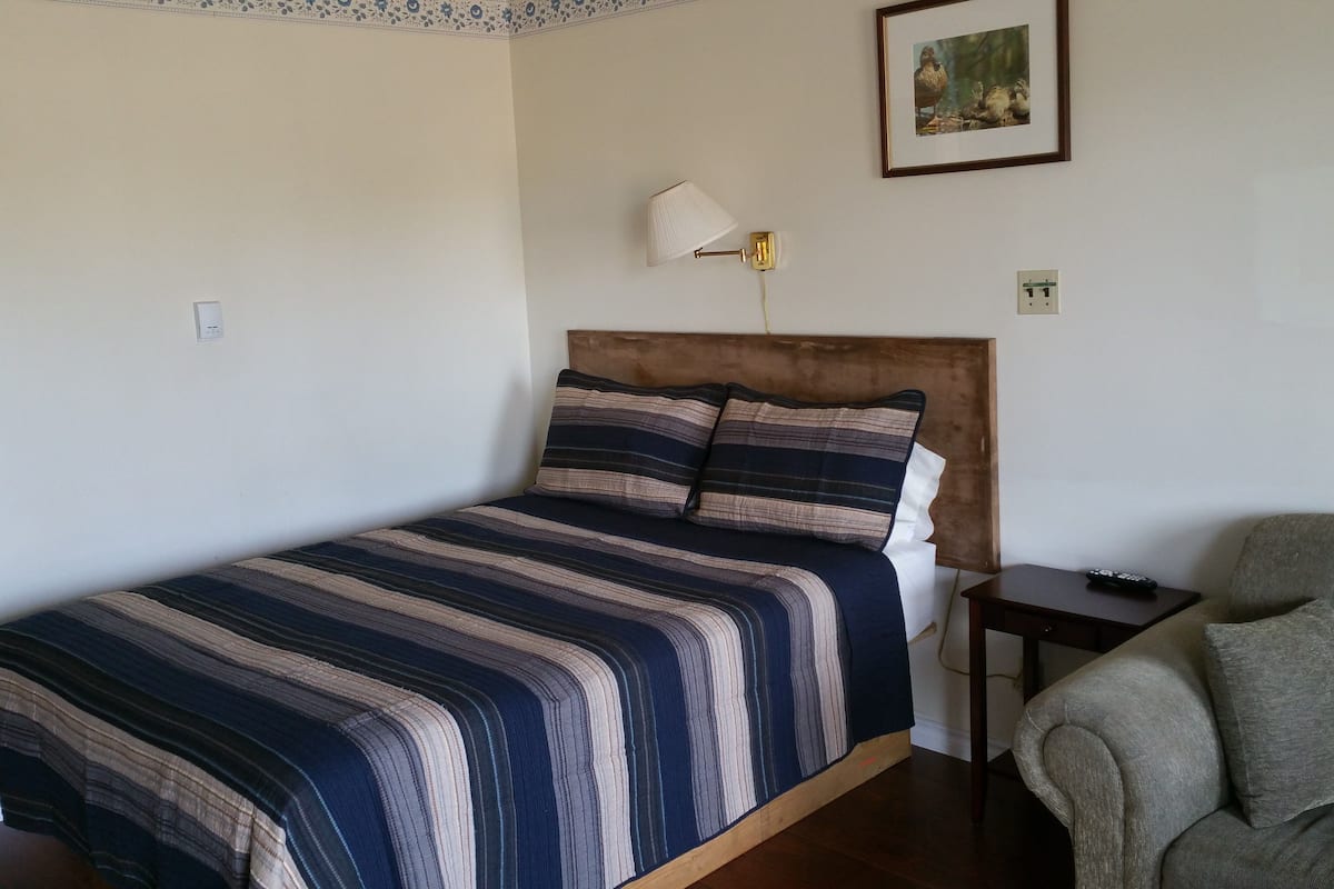A neatly made bed with striped bedding, a wooden headboard, a bedside table with a lamp, and a grey armchair.