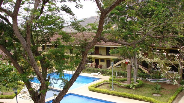 Uma área de piscina com duas piscinas retangulares, cercada por vegetação bem cuidada e um edifício com telhado telhado ao fundo.