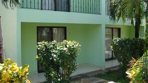 Un edificio de dos pisos con fachada verde, un balcón en el piso superior y un pequeño jardín con diversas plantas.