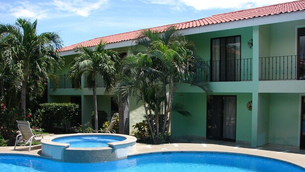 Una piscina con sillones de descanso, rodeada de palmeras y un edificio con techo de tejas rojas.