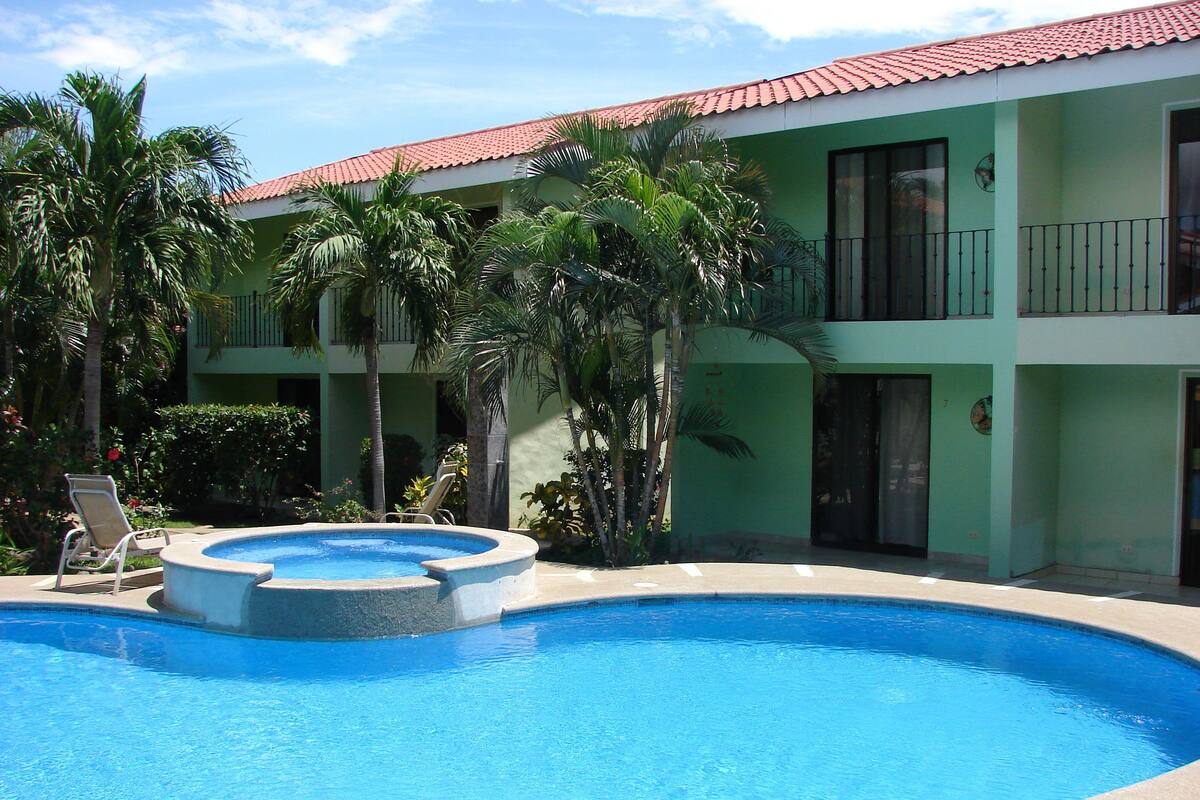 A swimming pool with lounge chairs, surrounded by palm trees and a building with a red-tiled roof.