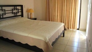 Una cama bien tendida con una colcha estampada, una mesita de noche con lámpara y una ventana con cortinas.