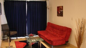 Una sala de estar con un sofá rojo, una mesa de centro de vidrio, un sillón, un tapete negro y un florero con ramas.