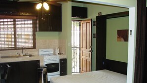 A compact kitchen with a stove, sink, and cabinets, a small trash bin, and a bed with a mattress. - Las Torres Studio (Sardinal)
