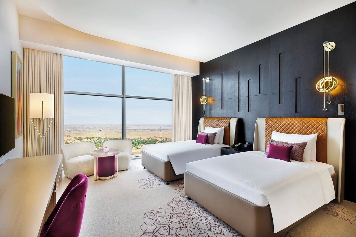 A hotel room with two beds, a desk, a chair, a lamp, and a view of the desert.
