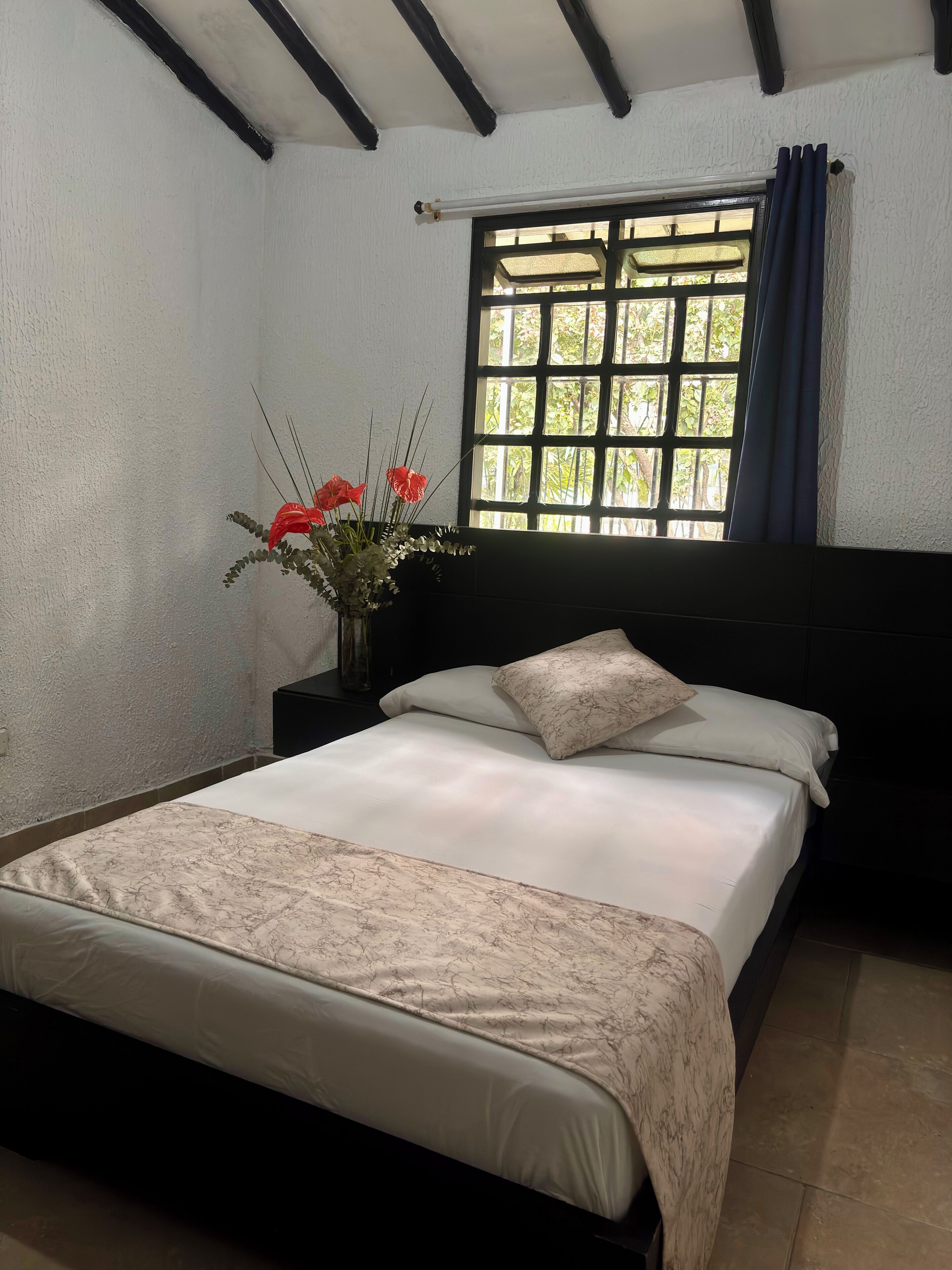 Un dormitorio con cama, mesita de noche con un florero, ventana con cortinas y pared con textura.