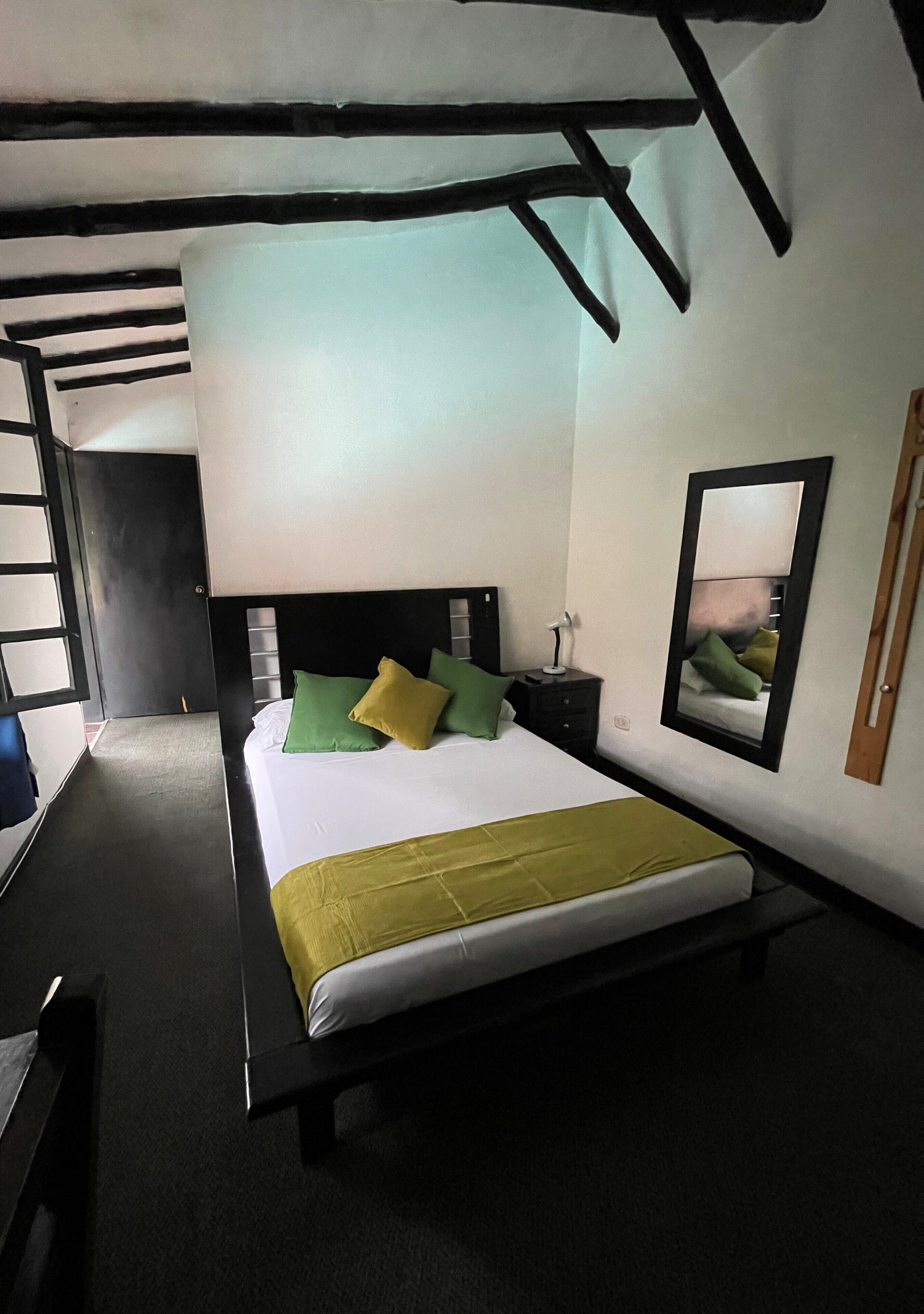 Een enkel bed met een witte matras, groene en gele kussens en een groene plaid. Er staat een nachtkastje met een lamp bij, en er hangt een spiegel aan de muur. De kamer heeft een zwart hoofdbord en een zwart bankje aan het voeteneinde van het bed.