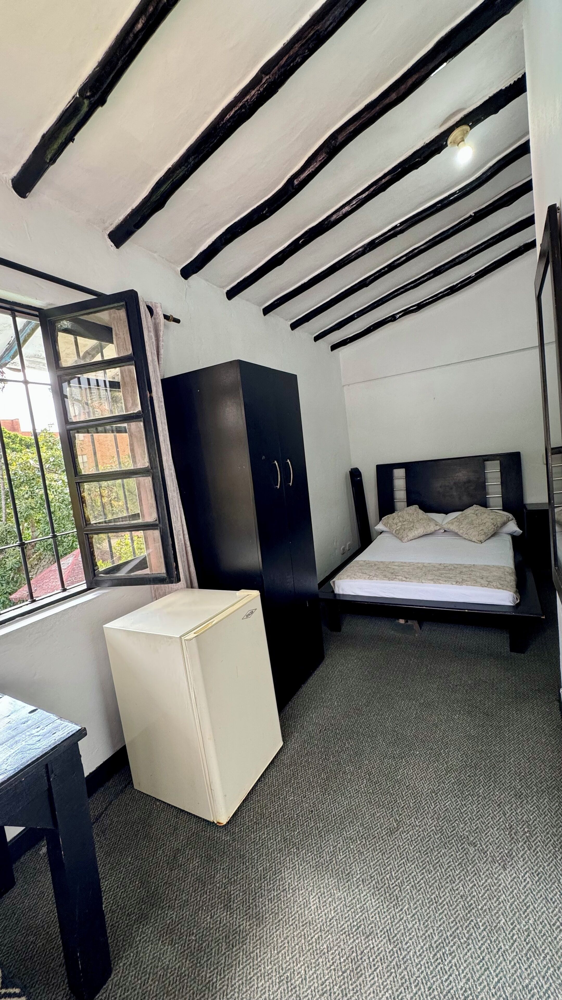A bedroom with a bed, a wardrobe, a small table, and a window with a view of greenery.