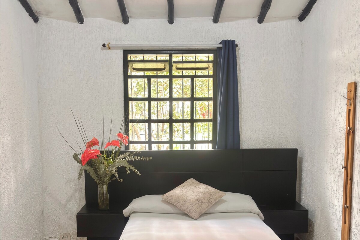 A bedroom with a bed, a nightstand, a window with curtains, and a wall-mounted wooden rack.