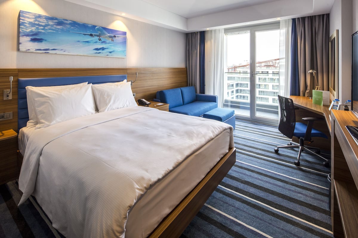A hotel room with a large bed, a desk with a chair, a sofa, and a view of the city through the window.