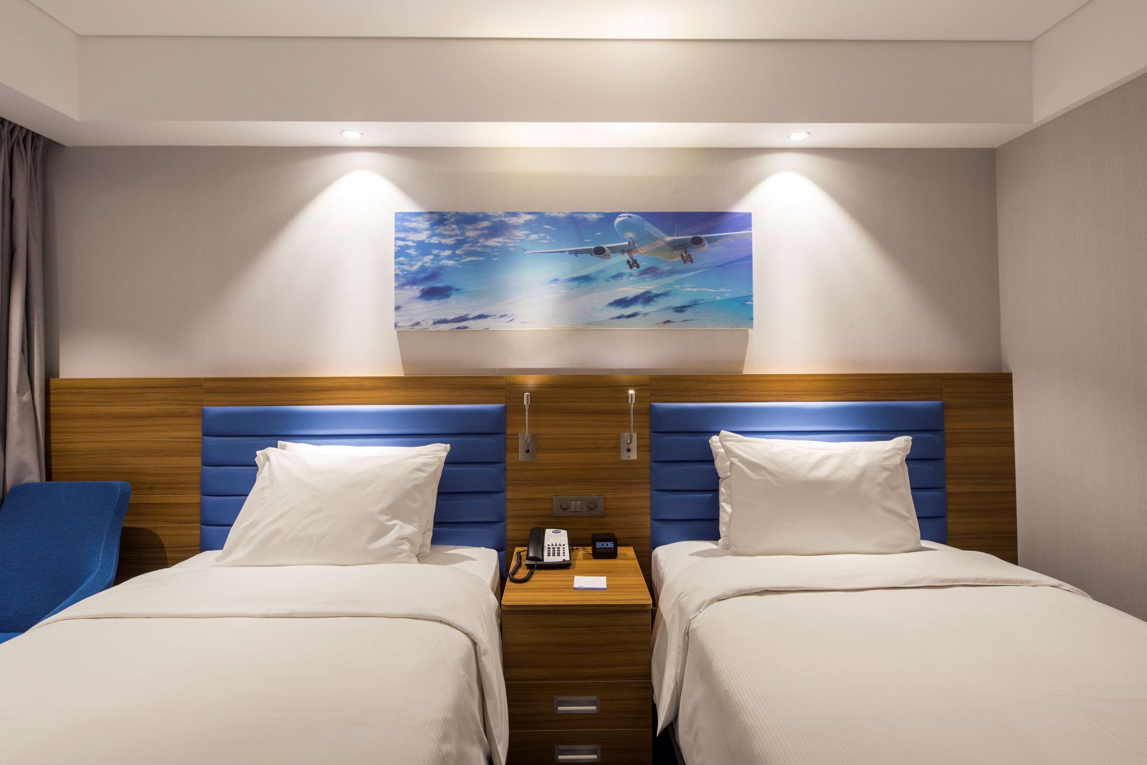 A hotel room with two beds, a wooden headboard, a nightstand with a phone, and a painting of an airplane.