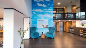 A modern lobby with a blue sky mural, two blue armchairs, a reception desk, and a vending machine. - Hampton by Hilton Istanbul Kurtkoy (Istanbul)