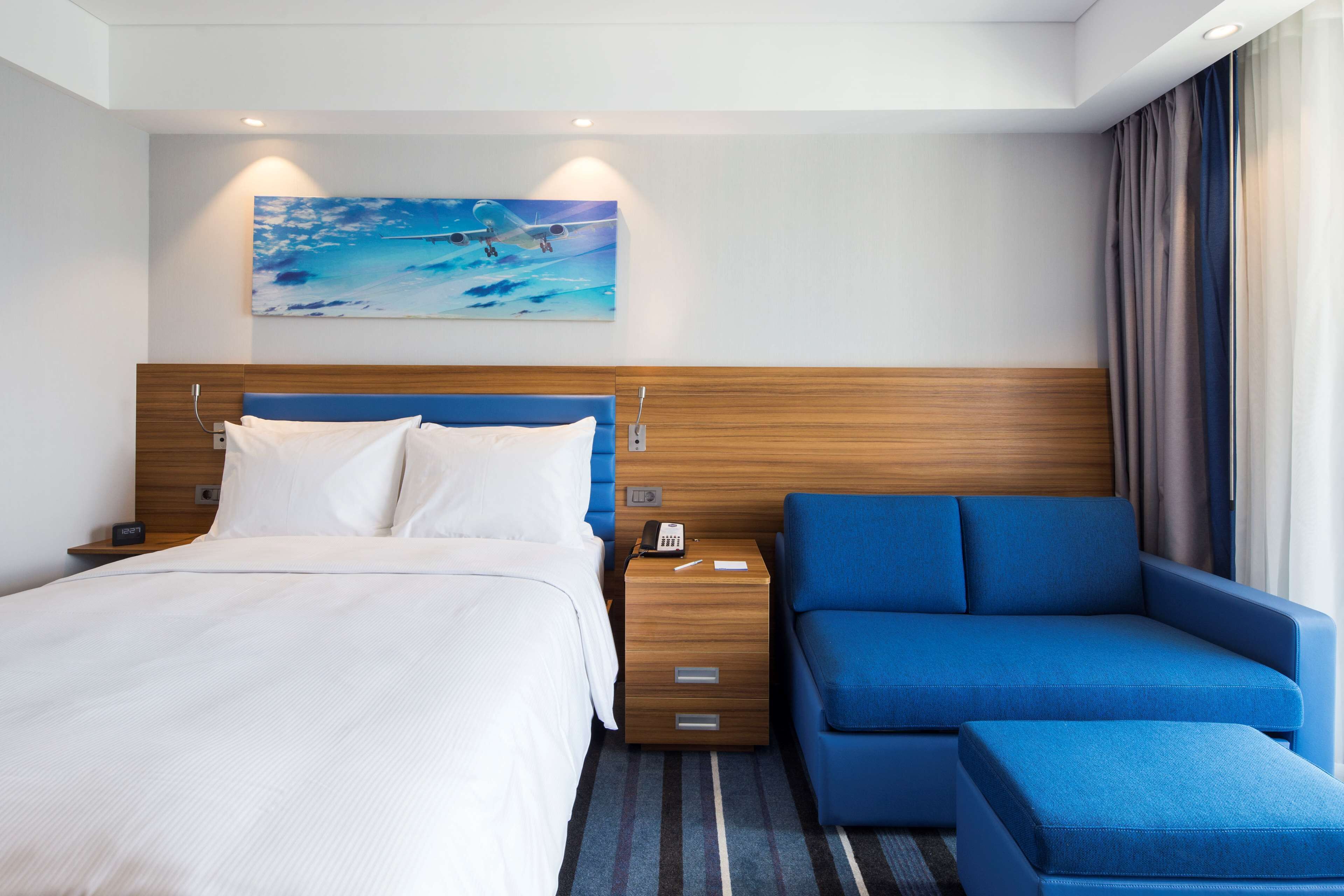 A hotel room with a bed, a blue sofa, a nightstand with a phone, and a painting of a beach scene.