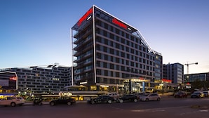A modern hotel with a prominent red sign, multiple floors, and a parking area with cars. - Hampton by Hilton Istanbul Kurtkoy (Istanbul)