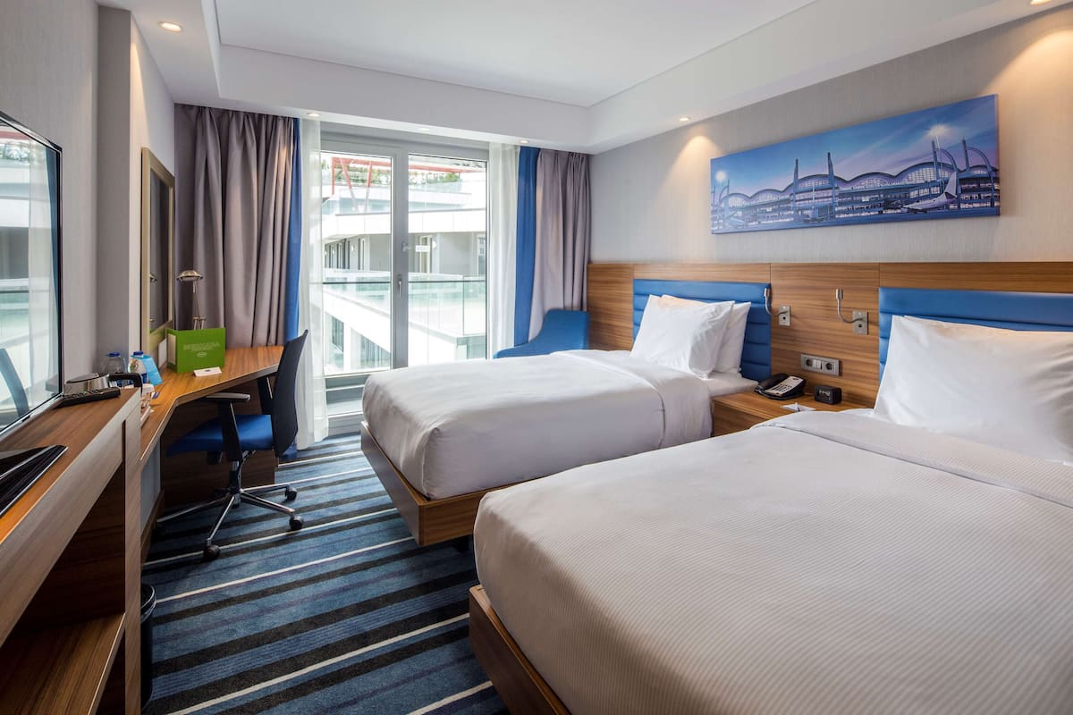 A hotel room with two beds, a desk, and a view of a building through the window.