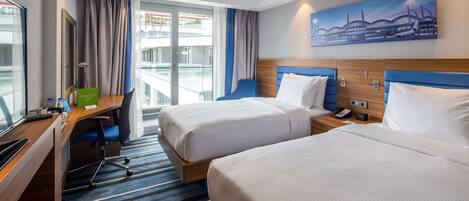 A hotel room with two beds, a desk, and a view of a building through the window.