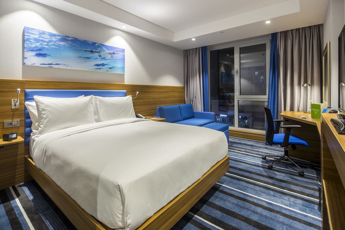 A hotel room with a large bed, a desk, a chair, a window with curtains, and a painting on the wall.