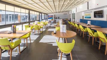 A modern cafeteria with long tables and bright yellow chairs, large windows, and artwork on the walls.