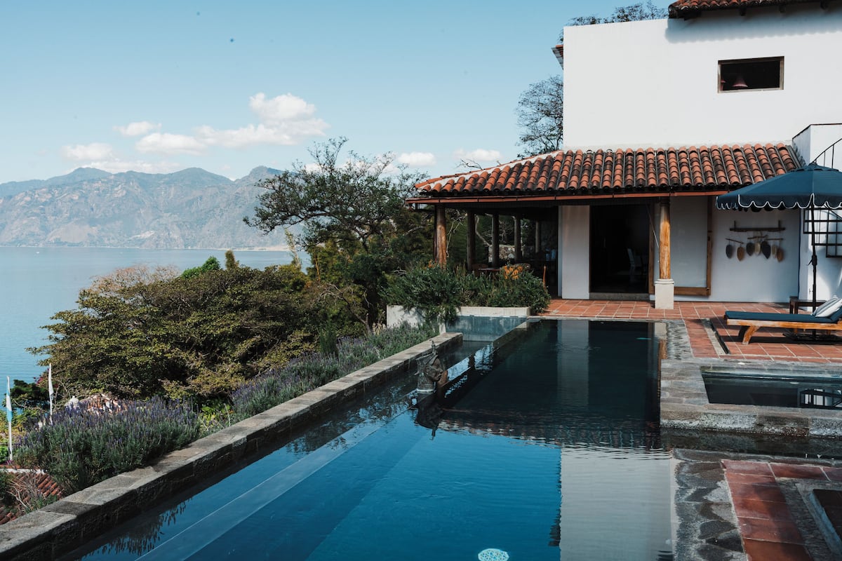Une piscine avec vue sur la mer, un bâtiment blanc avec un toit en tuiles et une terrasse en bois dotée de chaises longues.