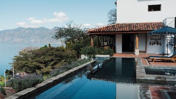 Una piscina con vista al mar, un edificio blanco con techo de tejas y una terraza de madera con sillones de descanso.