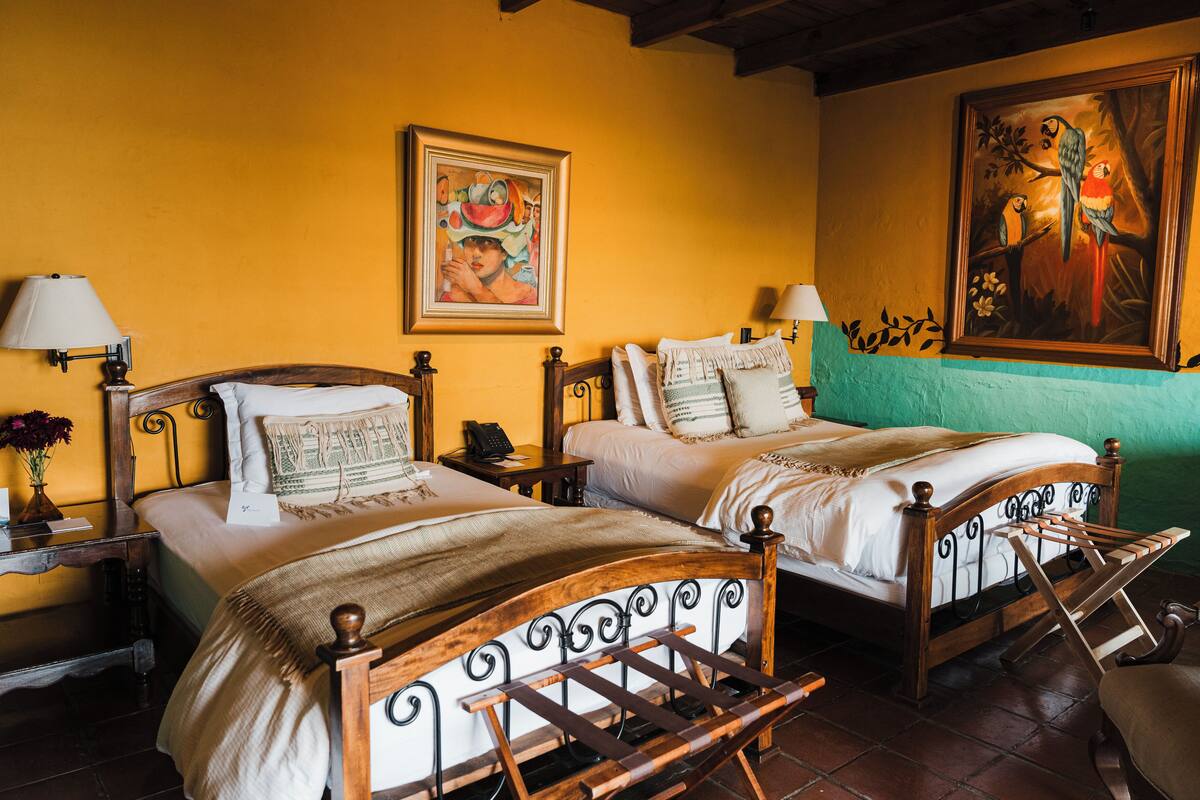Une chambre avec deux lits en bois, chacun doté d’une tête de lit et d’un pied de lit, flanqués de tables de chevet avec des lampes. Il y a un tableau au-dessus des lits et un mur vert sur le côté droit.