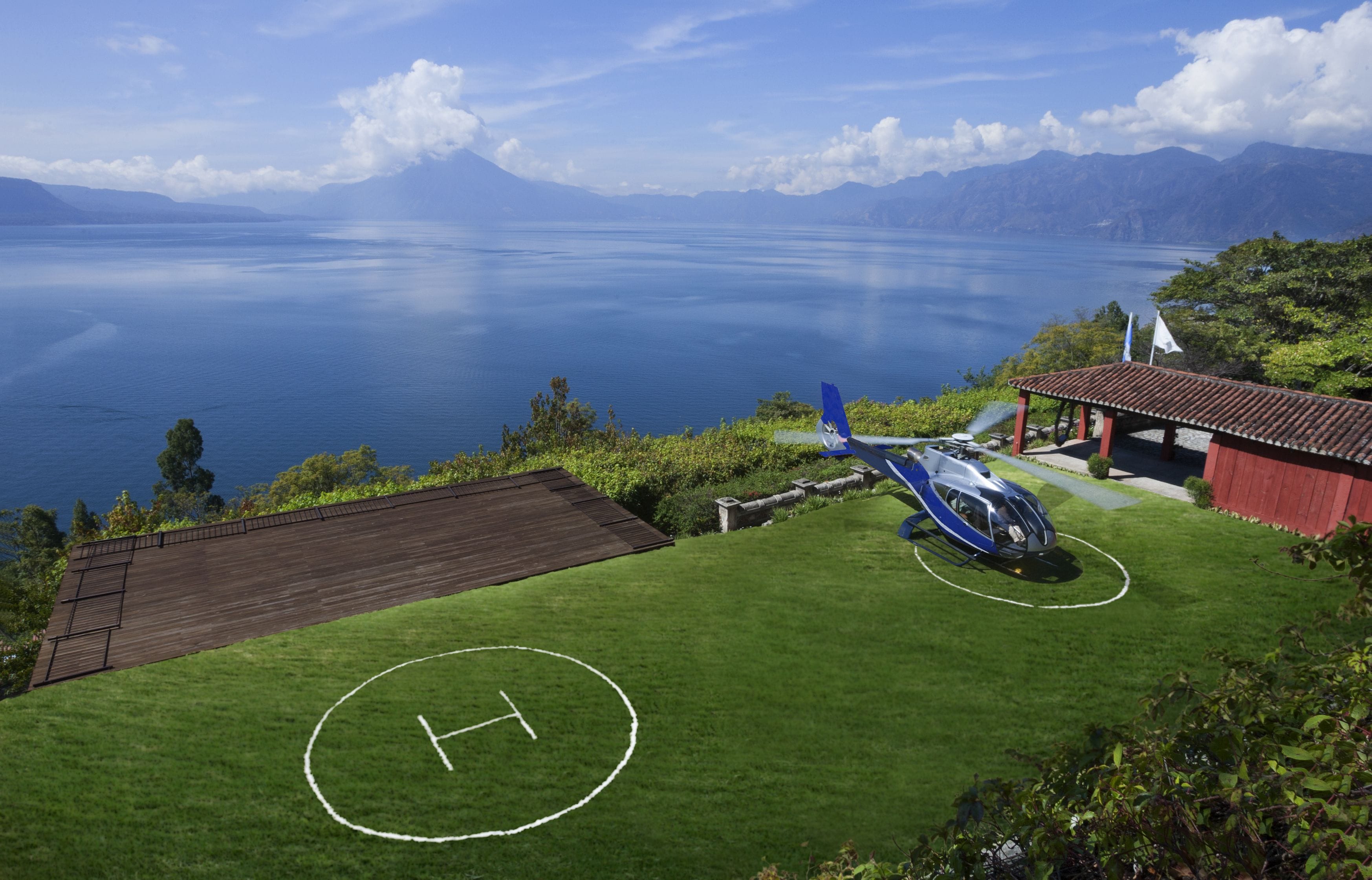 A helicopter on a grassy field with a helipad, a red building, and a lake with mountains in the background.