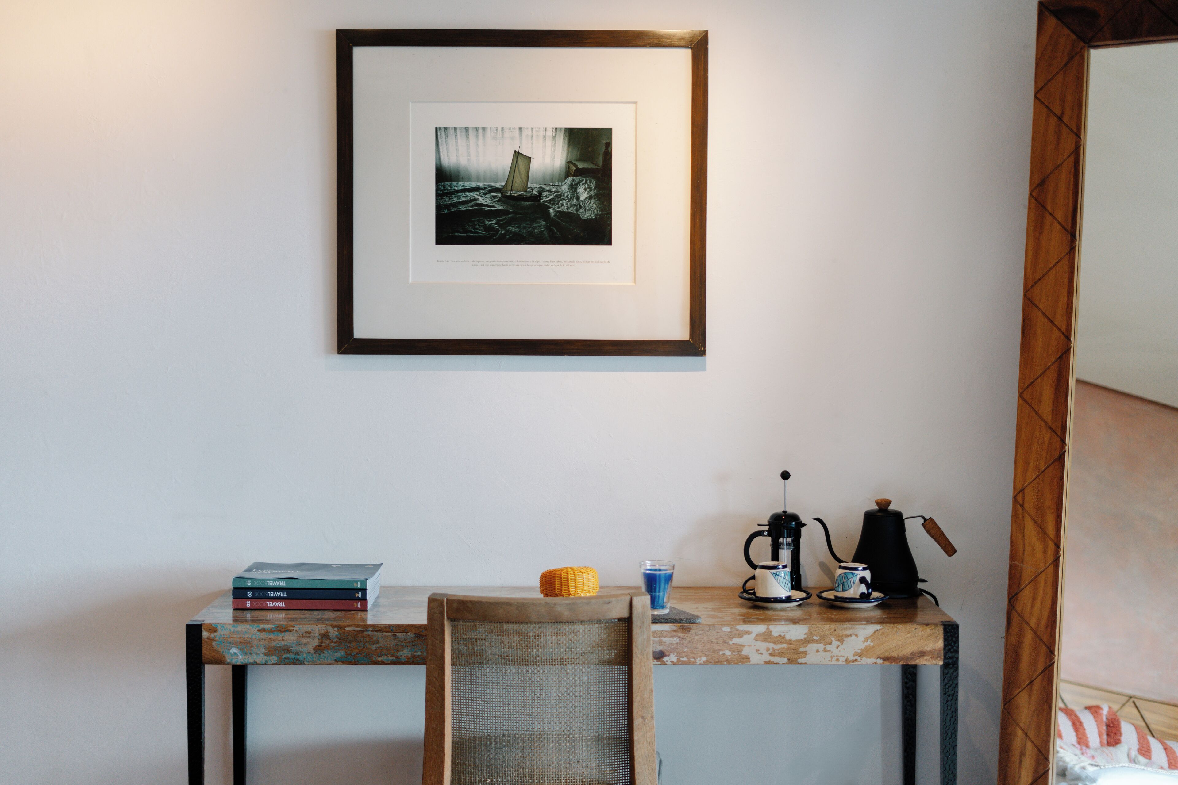 A room with a framed artwork on the wall, a small table with books, a chair, a coffee maker, and a cup.