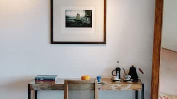 A room with a framed artwork on the wall, a small table with books, a chair, a coffee maker, and a cup.