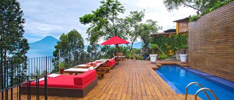 A wooden deck with red lounge chairs, a pool, and a view of a mountain.