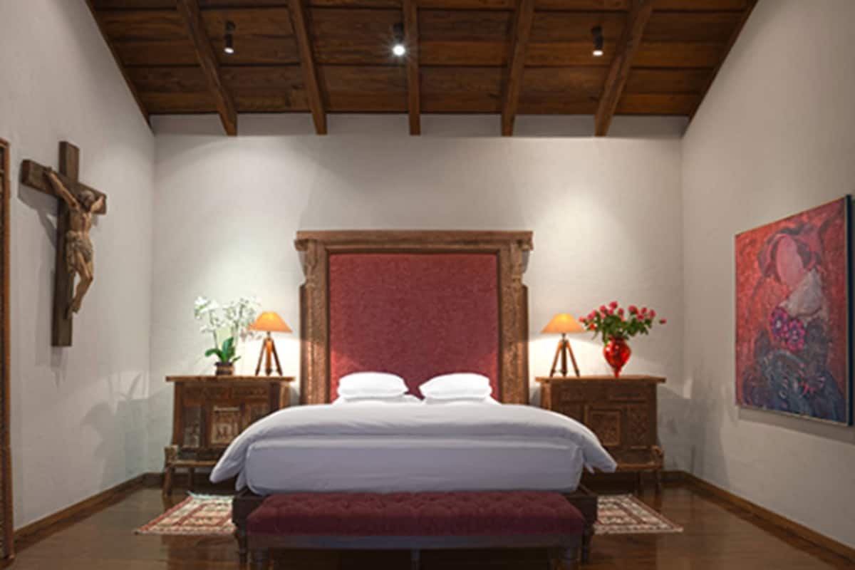 Une chambre avec un plafond en bois, un lit recouvert de draps blancs, un banc, deux tables de chevet avec des lampes et un tableau accroché au mur.
