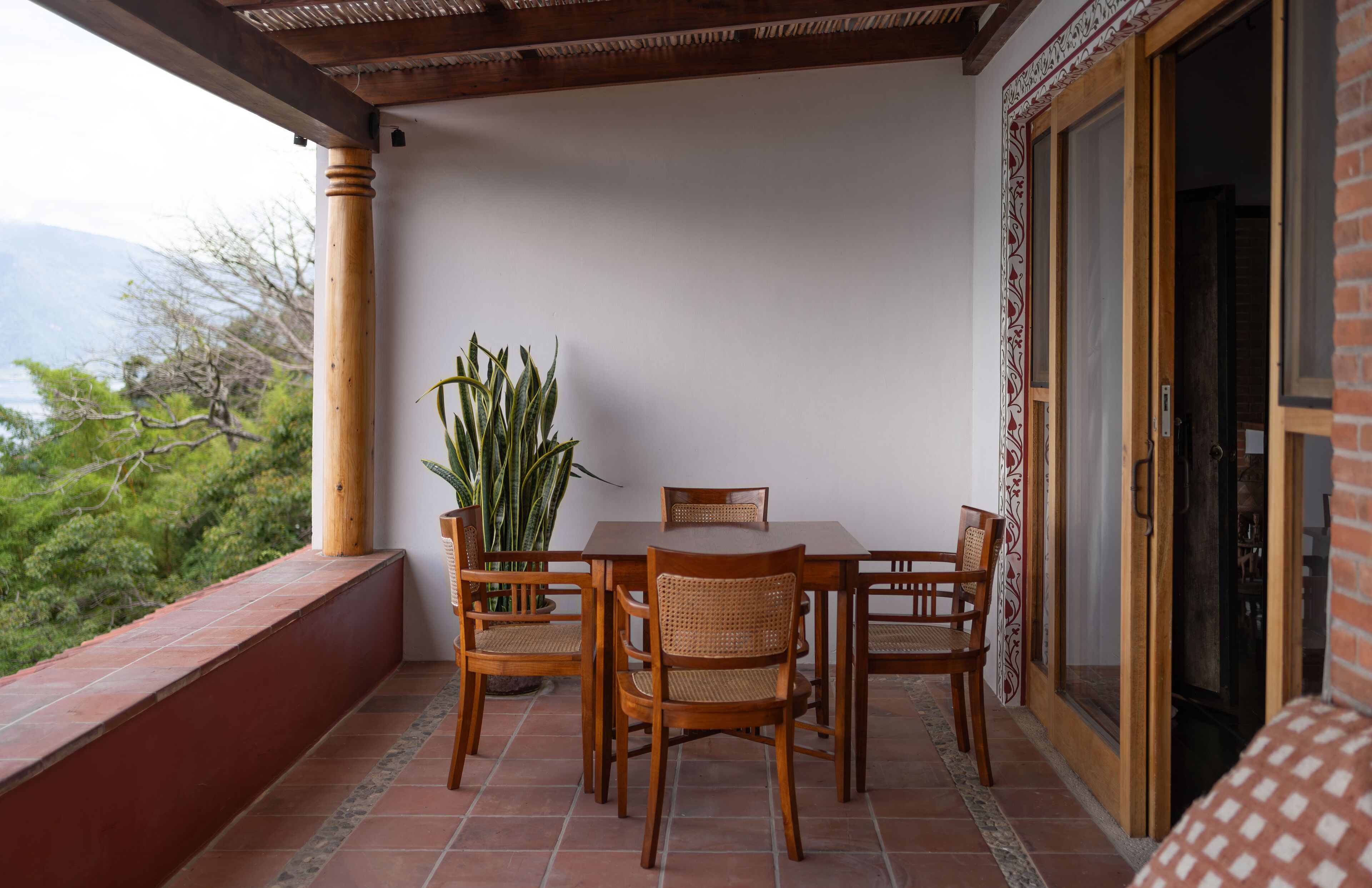 A wooden dining table with four chairs and a potted plant on a balcony with a tiled floor.