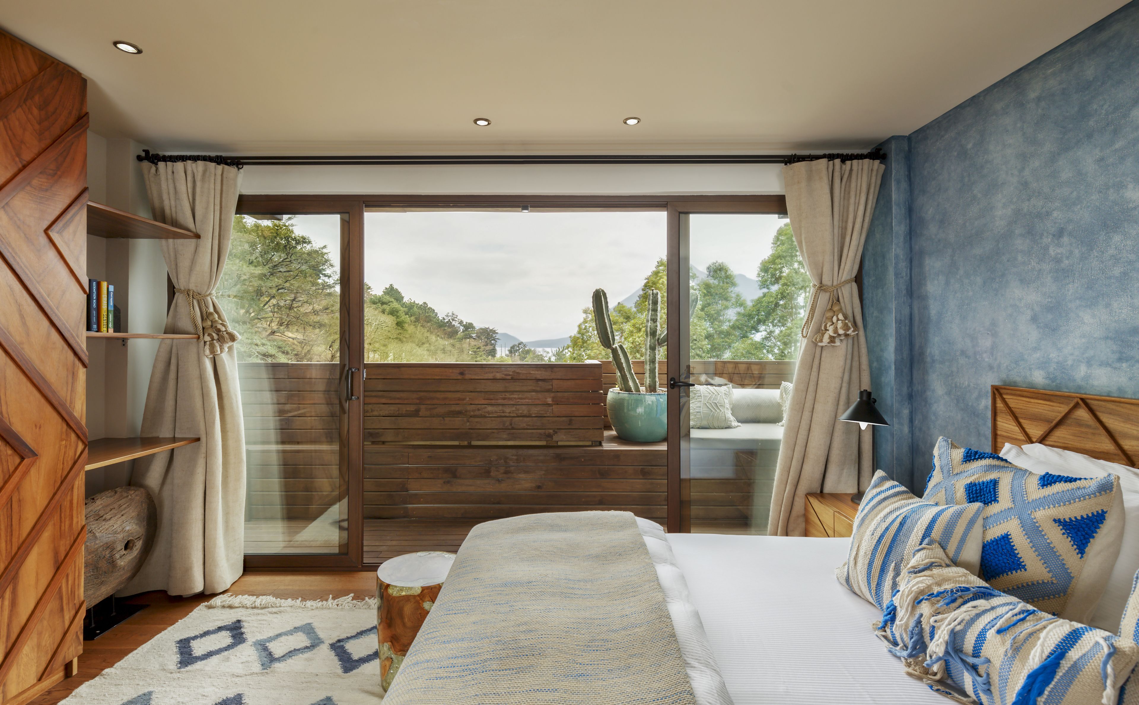 Un dormitorio con cama, cabecera de madera, ventana grande con cortinas, banco de madera y una planta de cactus.