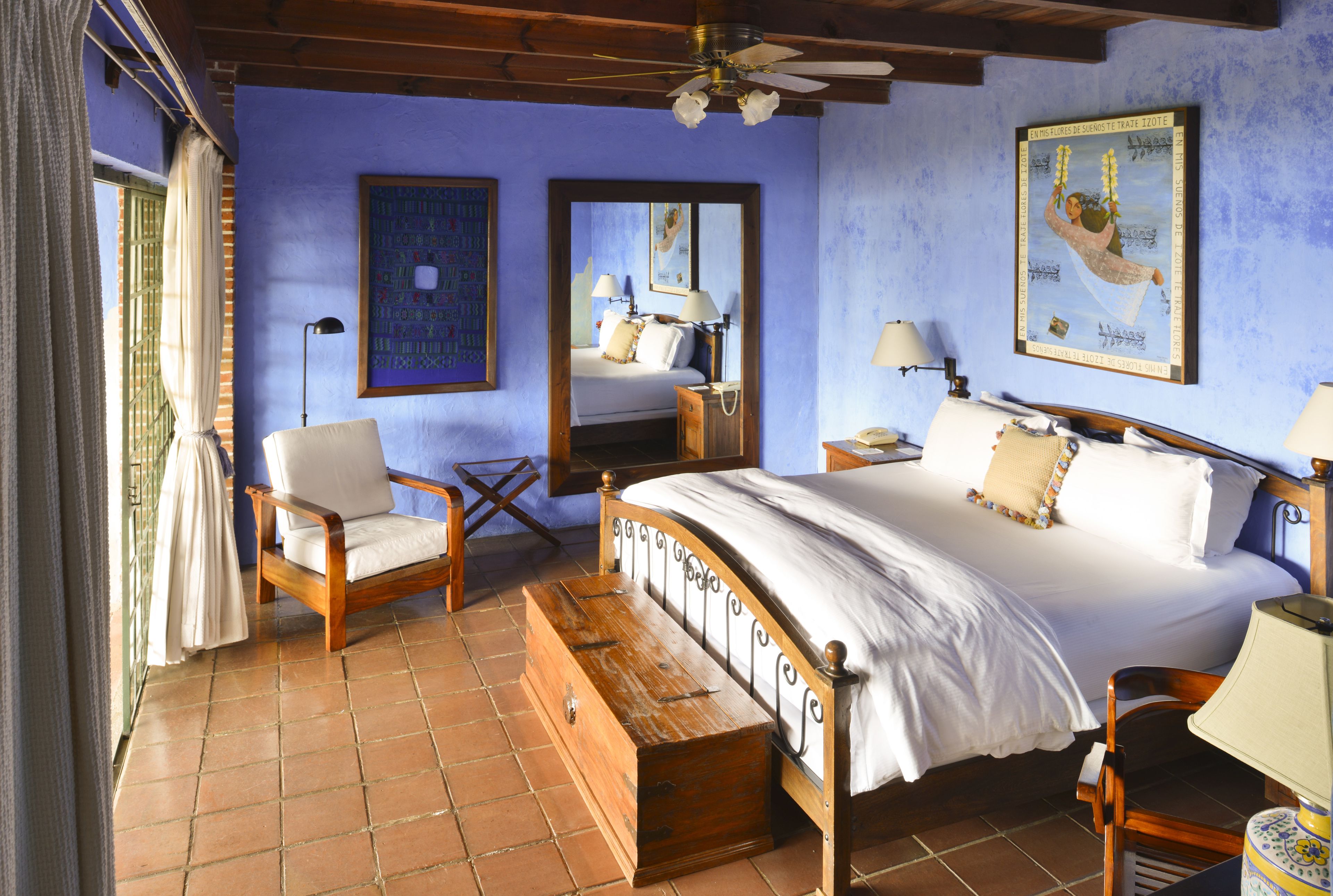 A bedroom with a bed, a wooden chest, a chair, a nightstand, a lamp, and a mirror.
