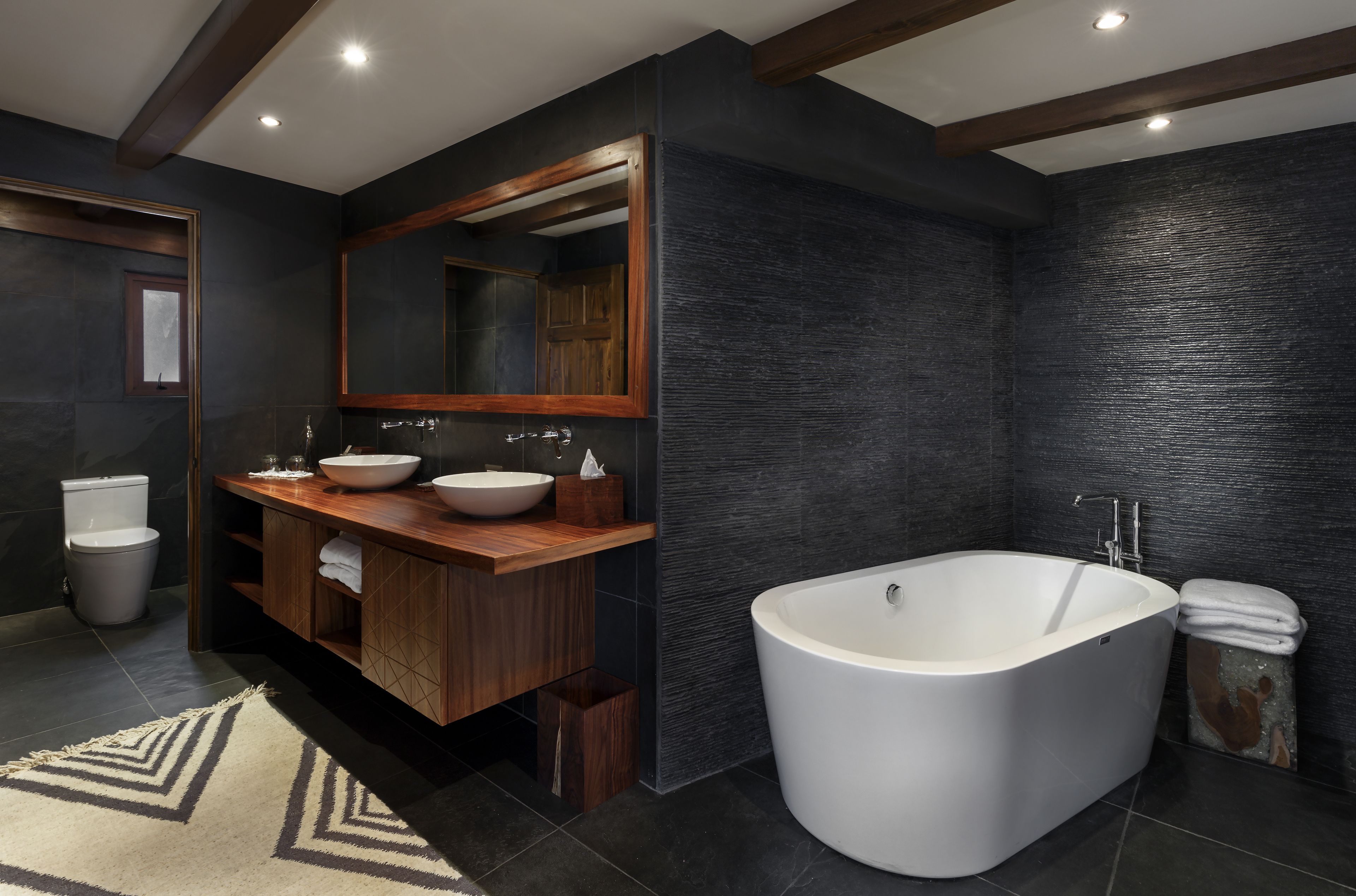 Un baño moderno con una bañera independiente, un tocador de madera con un espejo grande y una pared oscura con textura.