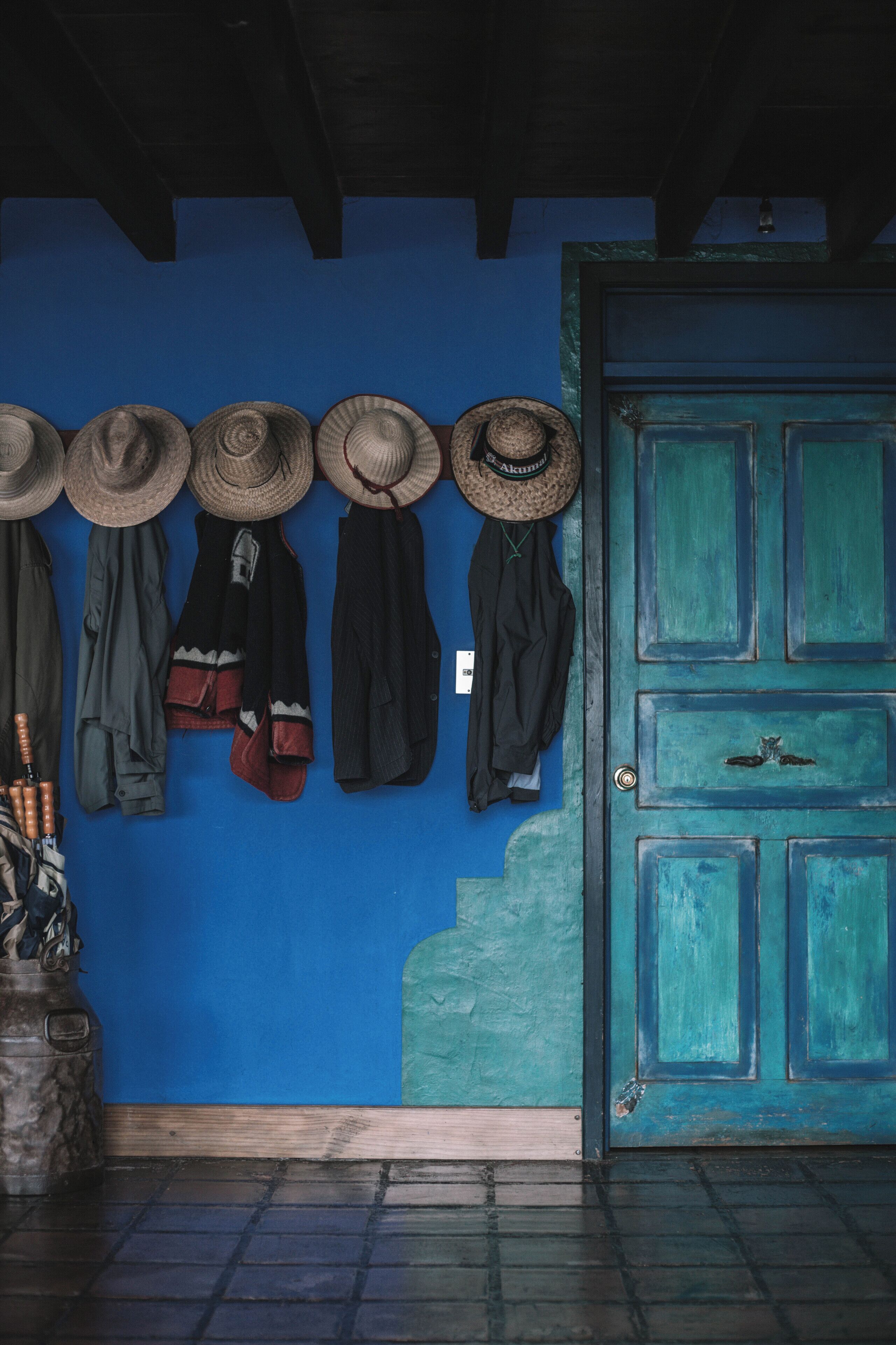 Pared azul con sombreros y abrigos colgados, una puerta verde y un suelo de baldosas.