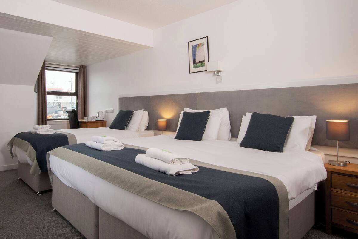 Una habitación de hotel con dos camas, una ventana con cortinas, una mesita de noche con lámpara y un cuadro en la pared.