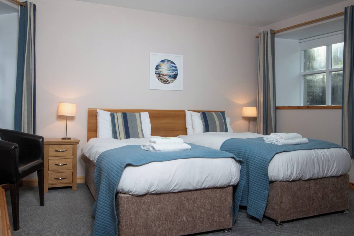 Habitación de hotel con dos camas, un escritorio y una silla. Se observa un cuadro en la pared y una ventana con cortinas.