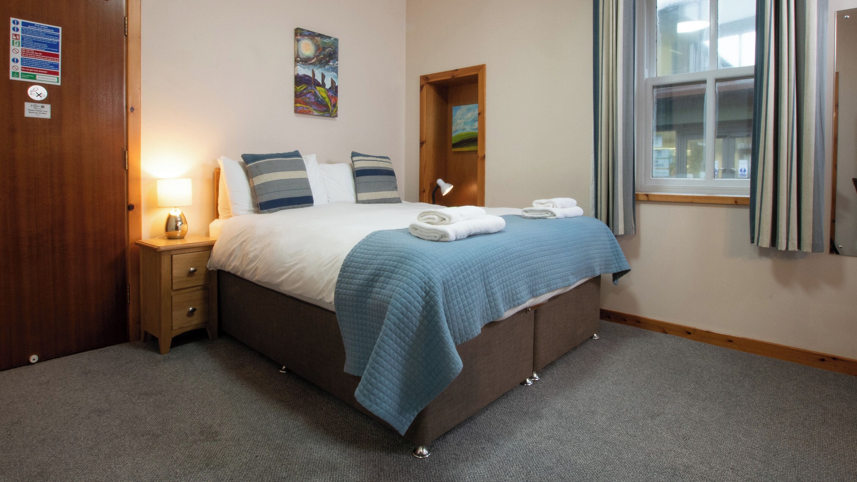 A neatly made bed with a blue blanket and pillows, a bedside lamp, and a window with curtains.