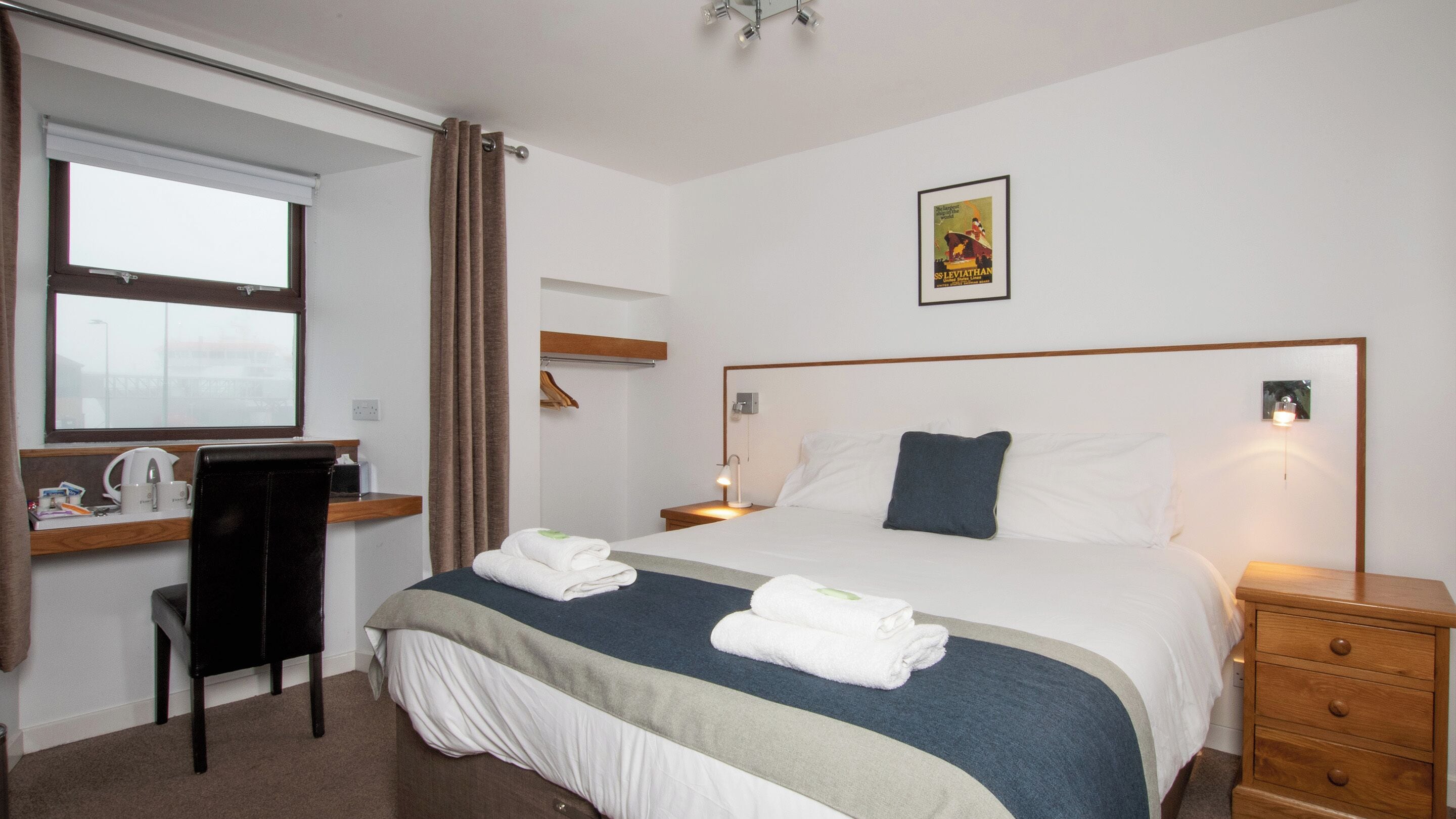 Habitación de hotel con cama, mesitas de noche, escritorio con silla, ventana con cortinas e imagen en la pared.
