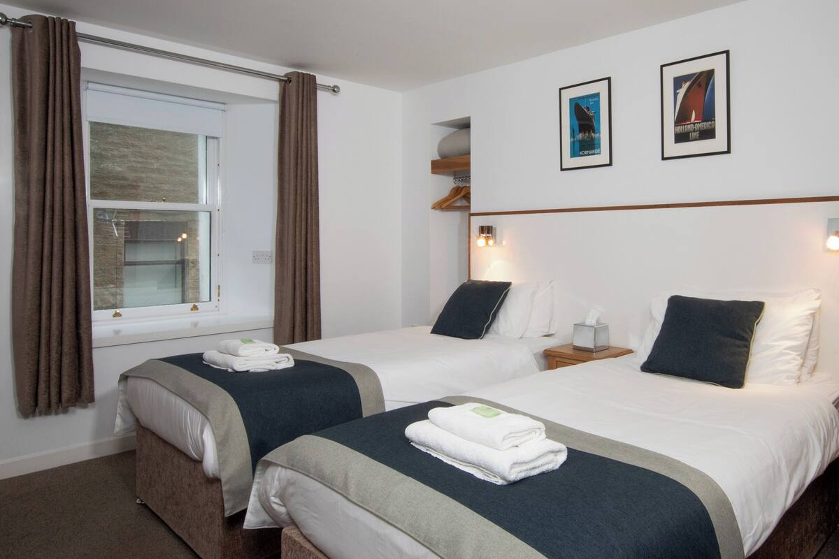 Habitación de hotel con dos camas, una ventana con cortinas, una mesita de noche con lámpara y dos cuadros en la pared.