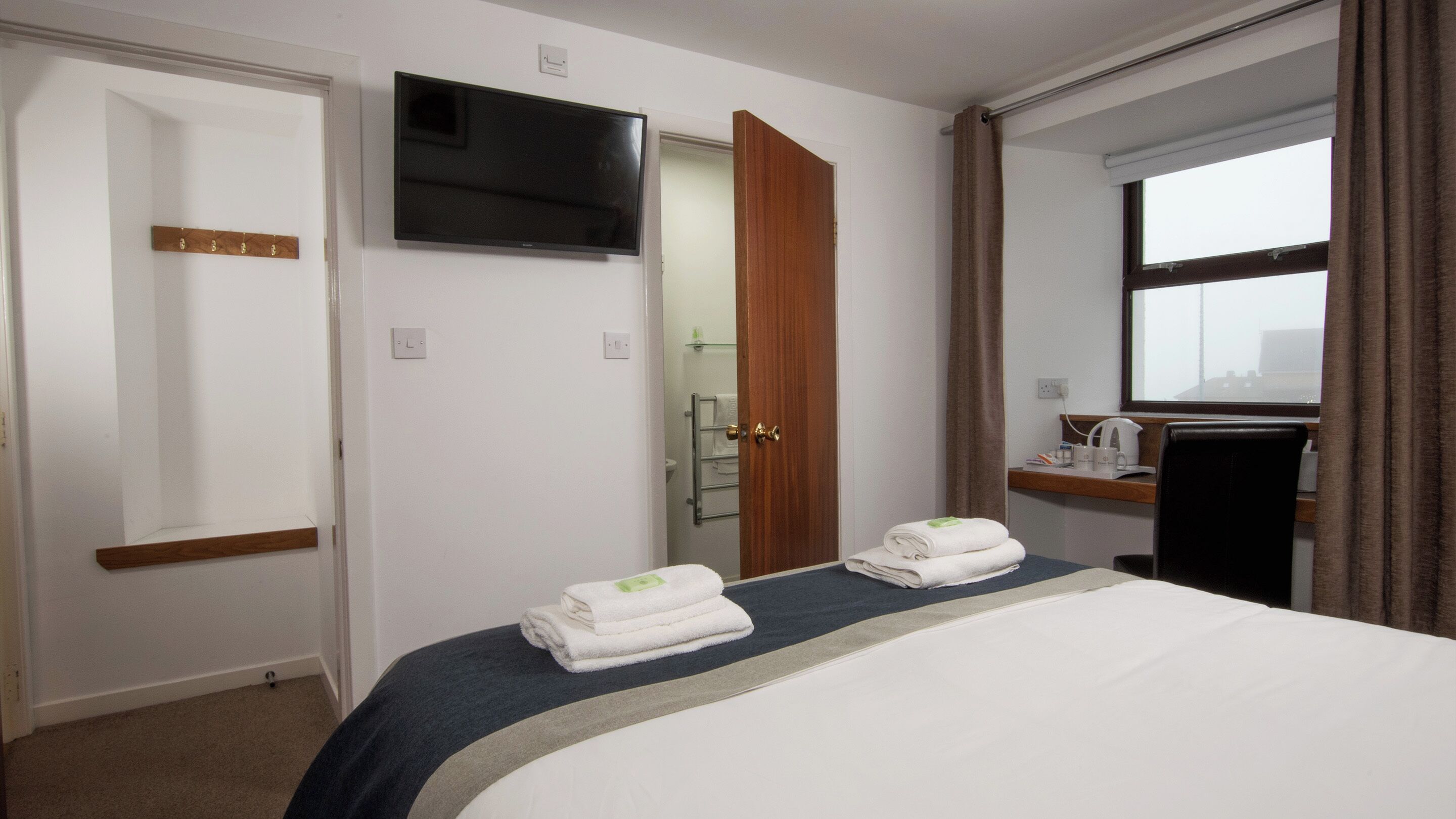A hotel room with a bed, a TV mounted on the wall, a bathroom visible through an open door, and a window with curtains.