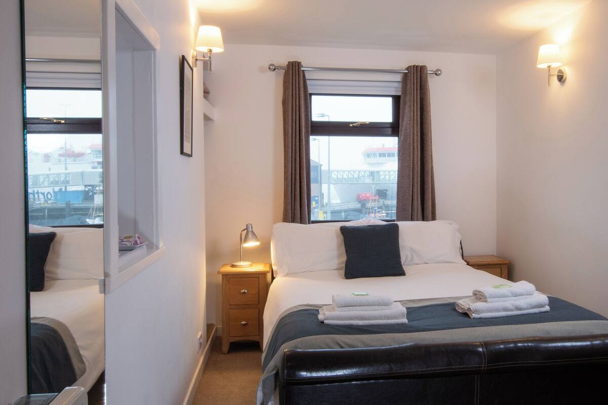 Habitación de hotel con cama, mesita de noche, lámpara, ventana con cortinas y vistas al exterior.