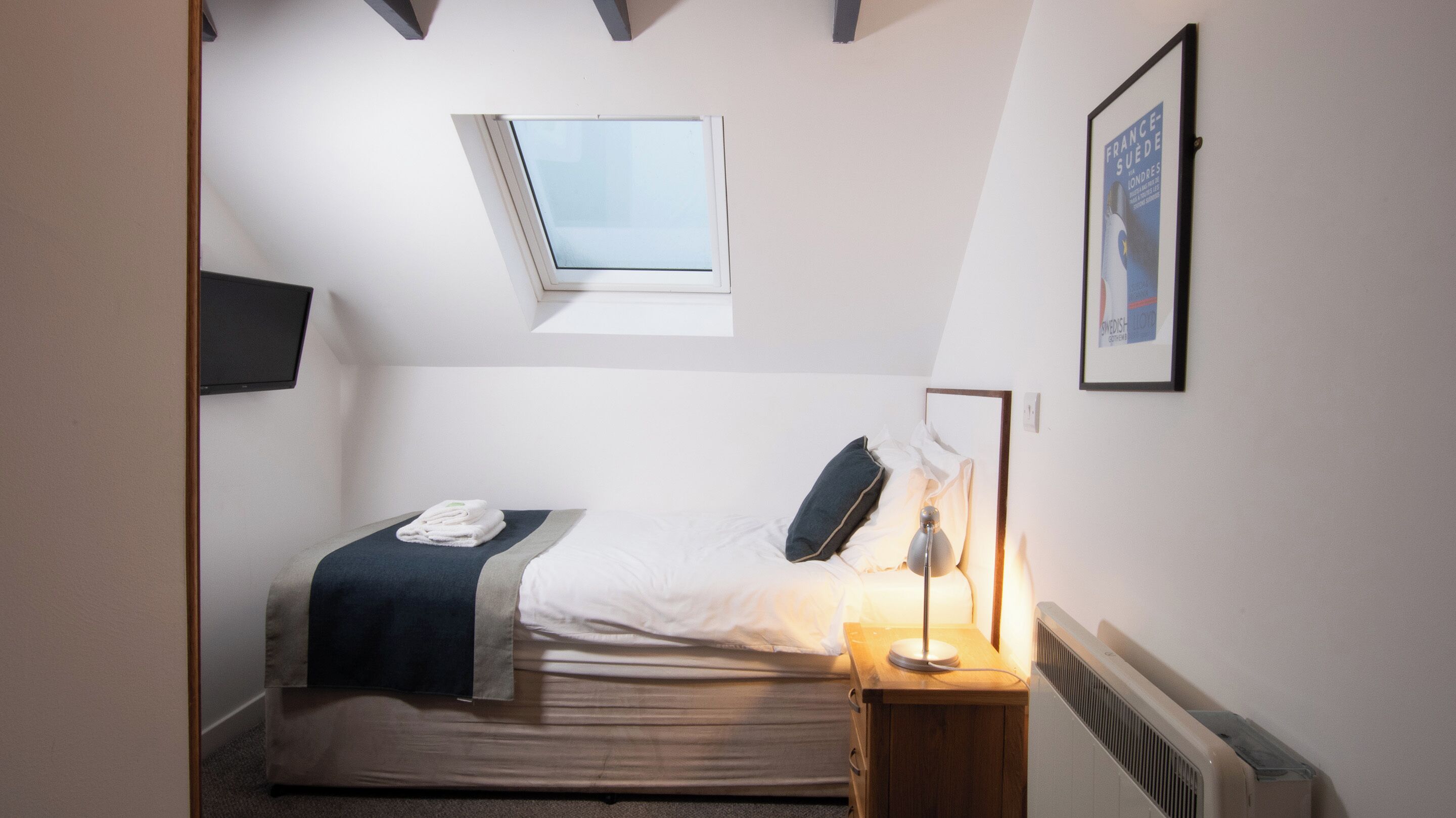 A small, neatly arranged bedroom with a single bed, a bedside table with a lamp, a radiator, a window, and a framed picture on the wall.