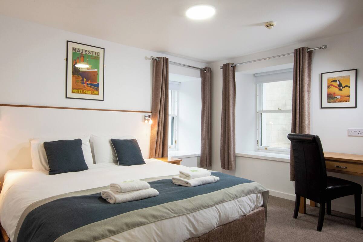 Una habitación de hotel con una cama grande, dos mesitas de noche, un escritorio con una silla y una ventana con cortinas.