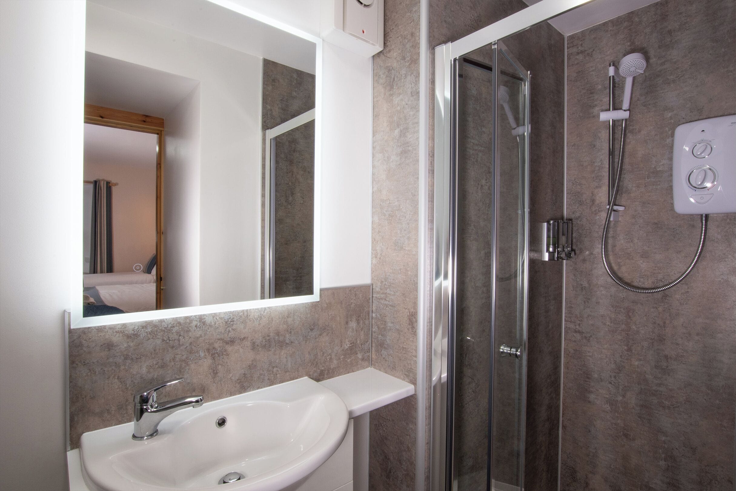 Un baño moderno con un lavabo blanco, cabina de ducha con mampara de vidrio y panel de control en la pared.
