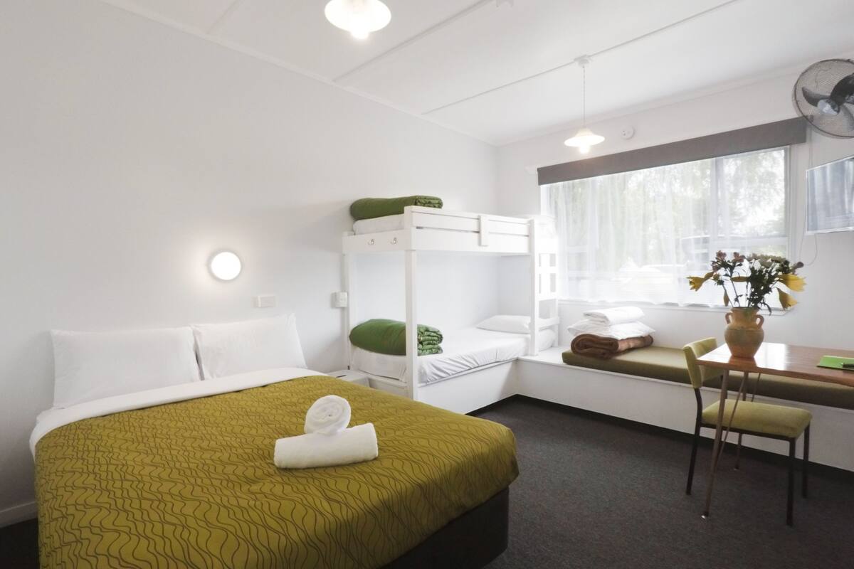 Een hotelkamer met een stapelbed, een bureau met stoel, een raam met gordijnen en een vaas met bloemen op het bureau.