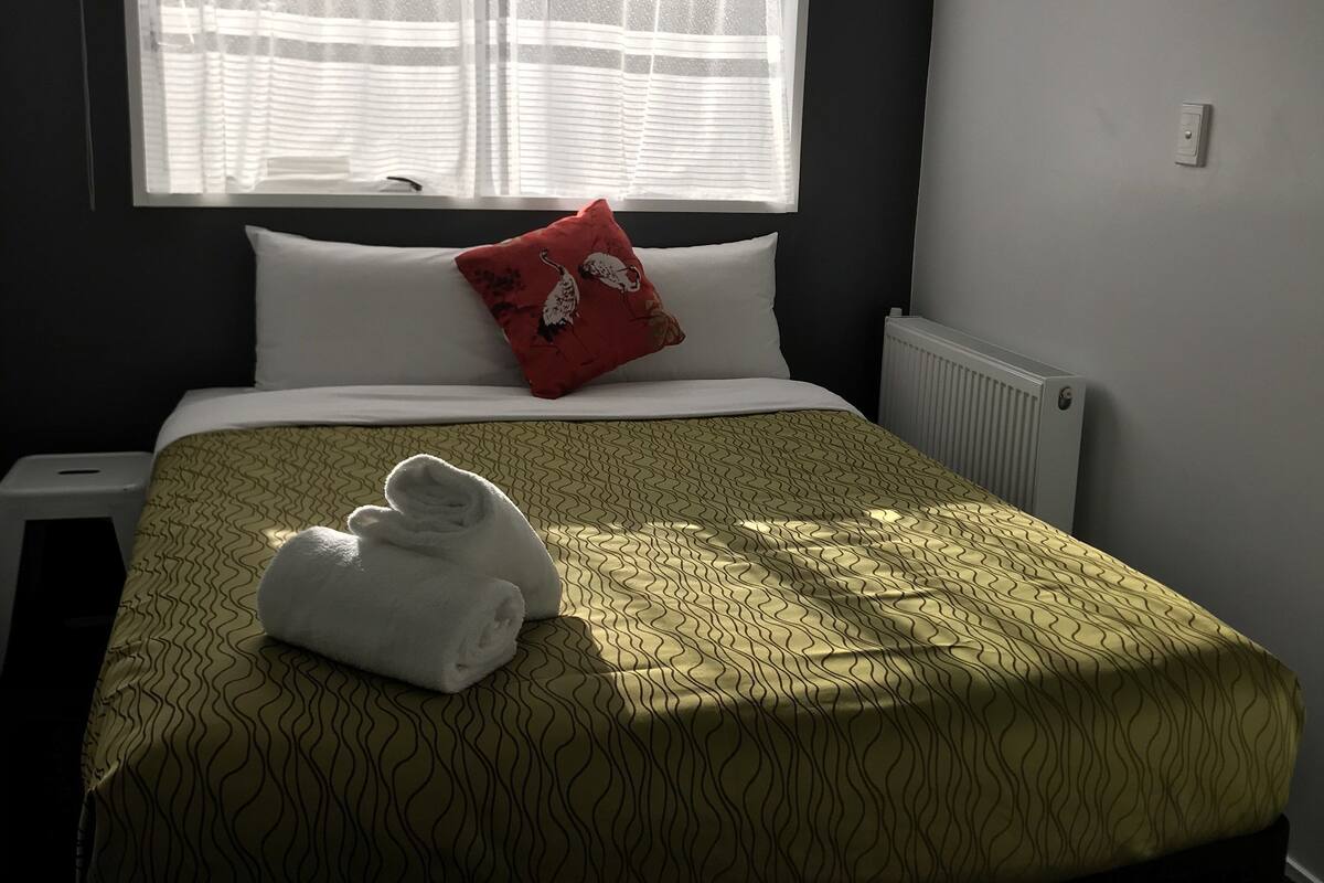 Een hotelkamer met een bed, een raam met dunne gordijnen, een rood kussen en een witte radiator.