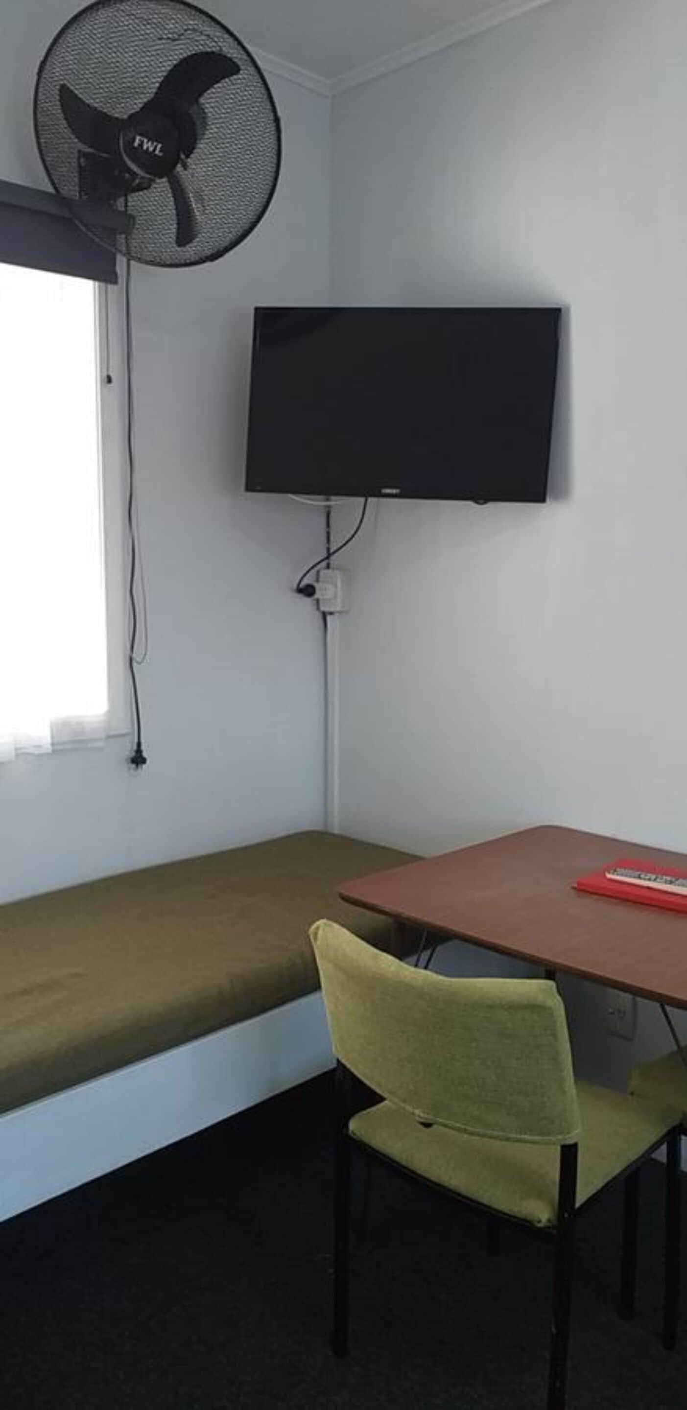Une chambre avec un lit, un bureau, une chaise, un téléviseur et un ventilateur sur pied.